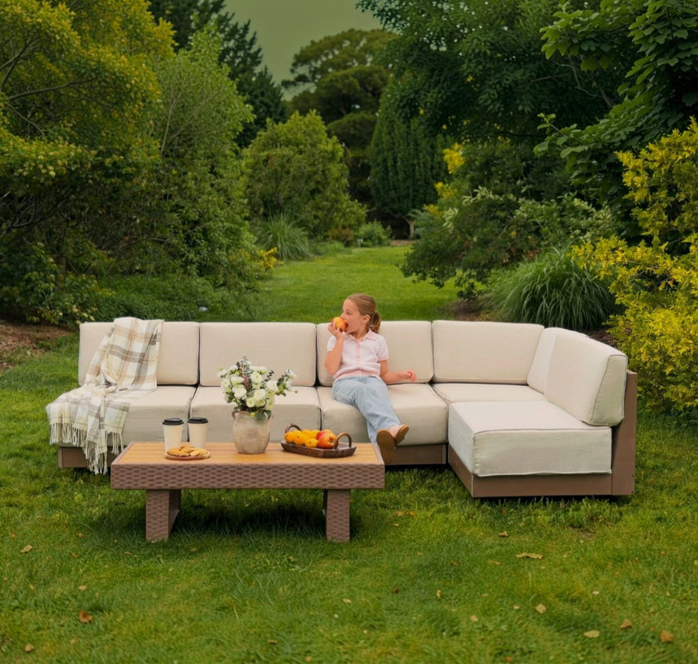 An outdoor sectional sofa with cream cushions and brown frames sits on a grassy lawn in a garden setting. A young girl is sitting on the sofa, and a matching throw blanket is draped over the left armrest.