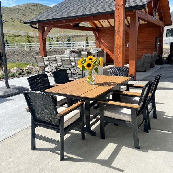 An outdoor dining set consisting of a rectangular wooden table and six matching armchairs with black woven backs and seats, cream cushions, and light wood arms, arranged on a concrete patio with a vase of sunflowers on the table.
