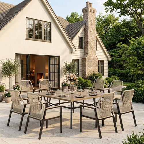 An 8-piece outdoor dining set with a rectangular wooden table and woven beige chairs, arranged on a stone patio in front of a white house with a stone chimney.