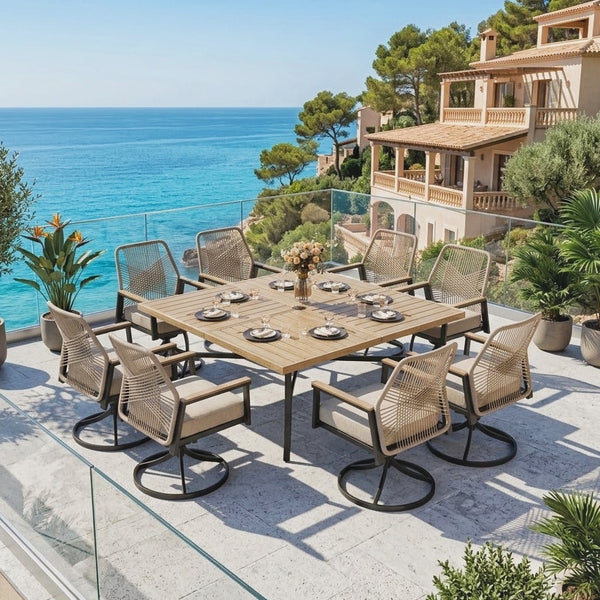 An outdoor dining set on a terrace overlooking the blue sea, featuring a rectangular wooden table and eight woven-back swivel chairs with beige cushions, set against a Mediterranean-style villa background.