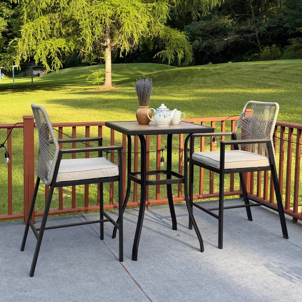 A three-piece outdoor patio bar set consisting of a square bar-height table and two matching barstools, shown on a concrete patio with a green lawn and trees in the background.