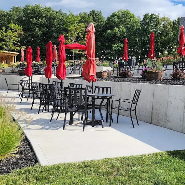 Outdoor seating area with tables, chairs, and red umbrellas in a garden setting.